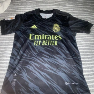 Real Madrid bortatröja Benzema - Säljer en svart Real Madrid bortatröja med gula detaljer och tryck. Tröjan har korta ärmar, Adidas-logga och klubbmärke på bröstet samt Benzema och nummer 9 på ryggen. Materialet är lätt och andas, perfekt för fotbollsträning eller match.
