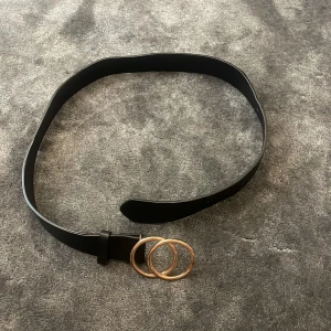 Svart bälte med dubbla guldringar - Stilrent svart bälte med två stora guldfärgade ringar som spänne. Bältet är i slätt material och har enkel design utan extra detaljer.