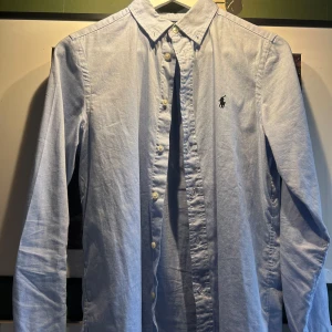 Ljusblå skjorta från Polo Ralph Lauren - Klassisk ljusblå skjorta från Polo Ralph Lauren med broderad logga på bröstet. Skjortan har  ärm, knappar framtill och är i slim fit-modell. Perfekt för en stilren look. 