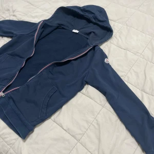 Mörkblå hoodie från Moncler - Säljer en mörkblå hoodie från Moncler med dragkedja och huva. Tröjan har långa ärmar, två fickor framtill och Moncler-logga på ärmen. Röd, vit och blå detalj längs dragkedjan ger en snygg touch.