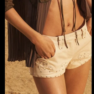 Beige shorts med broderi och nitar - Snygga beige shorts med vågformat broderi och små silverfärgade nitar längs kanterna. Modellen är kort och har bälteshällor samt knapp och dragkedja framtill. Perfekta för varma dagar på stranden eller festival. Helt nya!! Nypris 860kr