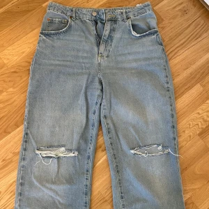 Ljusblå 90s straight jeans med slitningar - Säljer ett par ljusblå jeans i 90s straight modell med raka ben och slitna detaljer över knäna. Byxorna har klassisk femficksdesign, gylf med dragkedja och knapp samt medel hög midja. Perfekta för en avslappnad och trendig look.