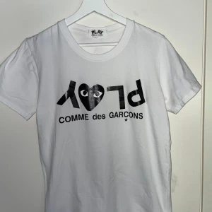 Vit t-shirt från Comme des Garçons Play - Vit t-shirt med svart tryck och det ikoniska hjärtögat från Comme des Garçons Play på bröstet. Klassisk rund hals och korta ärmar. Perfekt för en clean och stilren look.