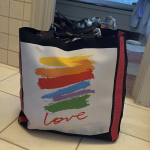 Stor shoppingväska från Tommy Hilfiger med regnbågstryck - Säljer en stor och rymlig shoppingväska från Tommy Hilfiger. Väskan är svart med breda röda band på sidorna och har ett färgglatt regnbågstryck samt texten 'love is bound words' på framsidan. Perfekt för att bära mycket! Helt ny! Ge gärna prisförslag!