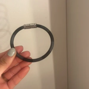 Svart Lv armband med magnetlås - Säljer ett äkta Louis Vuitton-armband i svart flätad design med silverfärgat spänne. Spännet är graverat med Louis Vuitton-loggan. Armbandet är i fint begagnat skick med mindre spår av användning. En stilren och tidlös accessoar som passar både till vardags och som present. Har varken kvitto eller förpackningen då jag haft den så länge och har slarvat bort det.