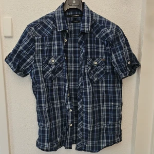 Blårutig kortärmad skjorta - Säljer en blårutig kortärmad skjorta med knappar framtill och två bröstfickor. Skjortan har klassisk krage och är perfekt för varma dagar. Snygg och avslappnad stil som passar till jeans eller shorts.