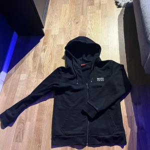 Svart hoodie från Hugo Boss - Säljer en svart hoodie från Hugo Boss med dragkedja och broderad logga på bröstet. Tröjan har huva och två fickor framtill. Perfekt för en avslappnad och stilren look.