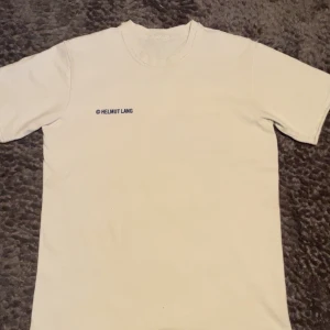 Vit t-shirt från Helmut Lang - Vit t-shirt från Helmut Lang med liten blå logga på bröstet och stor logga med texten 'VIENNA PARIS NEW YORK' på ryggen. Klassisk rund hals och normal passform. Perfekt för en clean och stilren look.