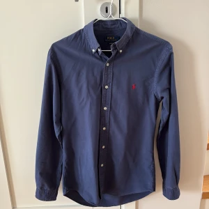 Mörkblå skjorta från Polo Ralph Lauren - Säljer en mörkblå skjorta från Polo Ralph Lauren i slim fit-modell. Skjortan har långa ärmar, knappar framtill och klassisk krage. Den ikoniska röda loggan är broderad på bröstet. Perfekt för dig som gillar stilrena plagg.