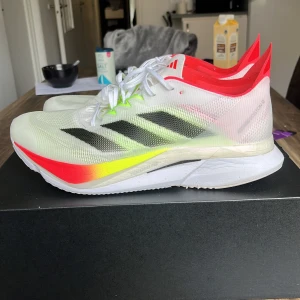 Adidas Adizero löparskor med neondetaljer - Säljer ett par Adidas Adizero löparskor i vitt med svarta ränder och neongula samt röda detaljer. Skorna har snörning och en lätt, ventilerande ovandel. Perfekta för dig som vill ha snygga och snabba sneakers till löpningen.