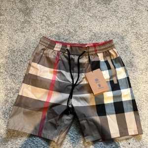 Rutigta Burberry shorts  - Säljer ett par klassiska Burberry shorts i deras ikoniska rutig mönster. Shortsen har elastisk midja med svart snörning. Perfekta för sommaren med snygg design och bekväm passform.