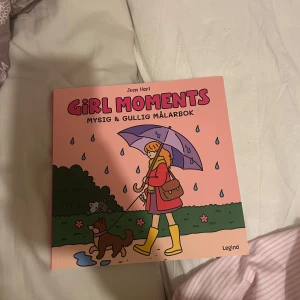 Girl Moments: Mysig & Gullig Målarbok - En söt och mysig målarbok av Jean Harl med rosa omslag. Boken innehåller olika sidor för att öva mönster, prova färger och fina illustrationer att färglägga, som en tjej med paraply och en katt. Perfekt för dig som gillar att pyssla och vara kreativ.