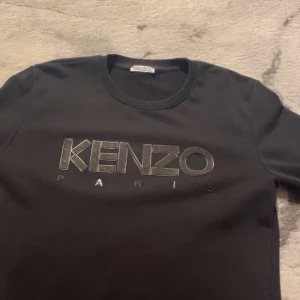 Svart långärmad tröja från Kenzo - Säljer en svart långärmad tröja från Kenzo med stort logotryck i silver på bröstet. Tröjan har rund halsringning och är i mjukt material, perfekt för en stilren look. Passar dig som gillar klassiska märken och enkel design.