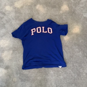 Blå t-shirt från Polo Ralph Lauren - Säljer en blå t-shirt från Polo Ralph Lauren med vit och röd POLO-text på bröstet. På ena ärmen finns en broderad amerikansk flagga och texten U.S.A. T-shirten har rund hals och är kortärmad. Perfekt för dig som gillar klassisk stil.