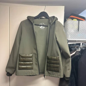 Olivgrön regnjacka från JOTT - Säljer en olivgrön jacka från JOTT med quiltade detaljer på fickor och huva. Jackan har dragkedja framtill, ribbade muddar och två fickor. Perfekt för regniga dagar och har en sportig look.