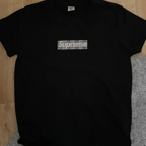 Svart Supreme x Burberry t-shirt - Svart t-shirt från Supreme och Burberry med klassisk Supreme box logo i Burberry-rutigt mönster på bröstet. Tillverkad i 100% bomull och har en normal passform. Perfekt för dig som gillar streetwear och exklusiva samarbeten.
