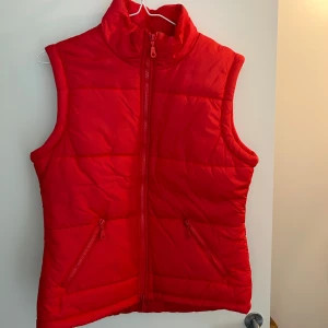 Röd dunväst / Fresh Spirit - Röd dunväst från Fresh Spirit med hög krage och dragkedja framtill. Västen har två fickor med dragkedja och quiltad design.