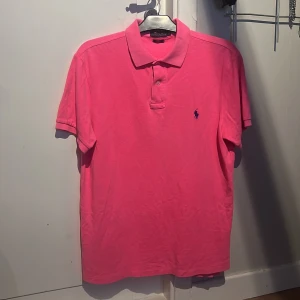 Rosa pikétröja från Polo Ralph Lauren - Säljer en klassisk rosa pikétröja från Polo Ralph Lauren med blå broderad logga på bröstet. Tröjan har korta ärmar, krage och knappar framtill. Perfekt för en stilren och sportig look.