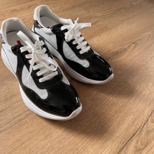 Svarta och vita sneakers från Prada - Säljer ett par snygga sneakers från Prada i svart och vitt. Skorna har vit mesh, svarta blanka partier och tydliga Prada-detaljer på plös och sula. Klassisk snörning och rund tå. Perfekta för dig som vill ha en stilren och exklusiv look.
