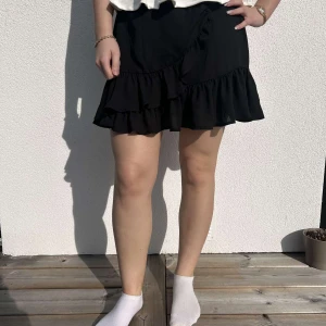 Svart Kjol från Gina Tricot - Snygg svart omlottkjol med volangdetaljer och knappar framtill. Perfekt för en stilren look. Kjolen har en kort längd och är lätt att matcha med olika toppar.