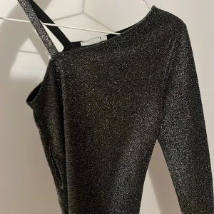 Svart glittrig one shoulder topp - Säljer en svart glittrig topp med one shoulder-design och en smal axelrem. Toppen har en åtsittande passform och är perfekt för att sticka ut. Materialet är stretchigt och skimrar i ljuset.