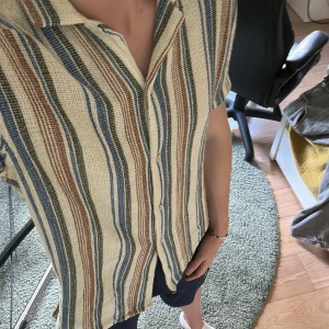 Randig skjorta i bomull med kort ärm från Zara  - Säljer en luftig skjorta med kort ärm och knappar framtill. Skjortan har ett randigt mönster i beige, blått, brunt och grönt. Perfekt för varma dagar och har en avslappnad passform. Storlek M 