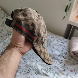 Beige keps från Gucci med monogram och röd-grön rand - Säljer en beige keps från Gucci med klassiskt GG-monogram och en ikonisk röd-grön rand på sidan. Kepsen har justerbart spänne bak och är tillverkad i bomullsmaterial. Perfekt för dig som gillar exklusiva accessoarer.