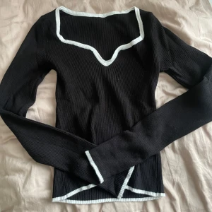 Svart ribbad långärmad topp med vit kant - Säljer en svart ribbad långärmad topp med vit kant längs halsringning, ärmslut och nederkant. Toppen har en unik v-ringad design och sitter snyggt på kroppen. Perfekt att styla med jeans eller kjol!