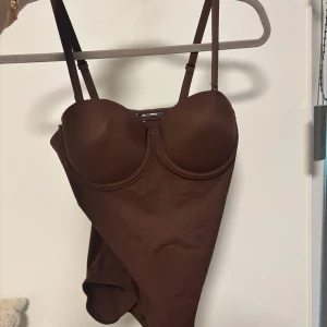 Brun body med justerbara band - Snygg brun body med vadderade kupor och bygel för extra stöd. Justerbara axelband och hyskknäppning i ryggen. Materialet är stretchigt och följsamt. Stolek S, helt oanvänd. Kan användas under kläder men även som en topp☺️