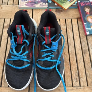 Adidas sneakers med blå och röda detaljer - Säljer ett par unika Adidas sneakers i svart mesh med blå snörning och röda ränder på sidan. Sulan har ett coolt mönster i blått, rött och vitt som verkligen sticker ut. Skorna har högt skaft och platt sula, perfekta för dig som vill ha något annorlunda.