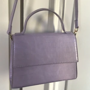 Ljuslila axelväska med krokoprägling - Snygg ljuslila axelväska med krokopräglat mönster och stilren design. Väskan har både axelrem och handtag, vilket gör den enkel att bära på olika sätt. Perfekt accessoar för att lyfta din outfit med en trendig touch.