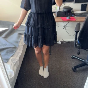 Svart kort volangklänning med öppen rygg - Supersöt svart kortklänning med volanger i flera lager och transparenta prickiga ärmar. Klänningen har öppen rygg med knytband och en luftig känsla, perfekt för varma dagar. Ärmarna är halvlånga och tyget är lätt och flowy. Aldrig använt då den är för stor 