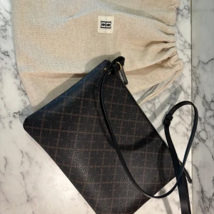 By Malene Birger Ivy axelväska - Snygg Ivy axelväska från By Malene Birger i mörkbrunt och svart med rutigt mönster. Väskan har axelrem, guldfärgade detaljer och dragkedja upptill. Tillverkad i tåligt syntetmaterial med exklusiv känsla.