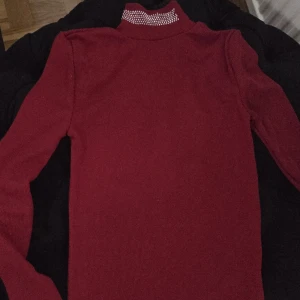 Vinröd polotröja med pärlor - Vinröd polotröja med lång ärm och snygga pärldetaljer på kragen. Tröjan är stickad och har en tight passform som sitter snyggt. Perfekt för dig som vill ha en stilren men ändå unik look.