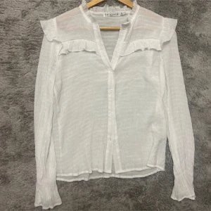 Vit blus med volang från By Clara - Säljer en vit blus från By Clara med långa ärmar och volangdetaljer vid axlar och krage. Blusen har knappar framtill och är tillverkad i ett lätt, krinklat bomullstyg som ger en luftig känsla. Perfekt för dig som gillar romantiska och feminina detaljer.