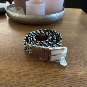 Svart och grått flätat bälte - Snyggt flätat bälte i svart, grått och vitt med detaljer i brunt skinn vid spännet. Bältet har ett klassiskt randigt mönster och ett silvrigt spänne. Perfekt accessoar för att lyfta din outfit med en sportig och stilren touch.