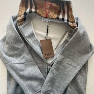 Grå zip-hoodie från Burberry, L men passar Som M - Grå hoodie från Burberry med dragkedja och klassiskt rutigt foder i huvan. Broderad Burberry-logga på bröstet och ribbade muddar.