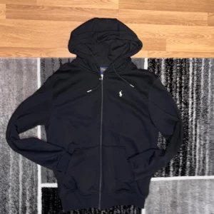 Svart hoodie från Polo Ralph Lauren - Svart hoodie från Polo Ralph Lauren med dragkedja och klassisk vit logga på bröstet. Tröjan har huva med snörning, två fickor fram och långa ärmar. Perfekt för en chill och stilren look. Aldrig använd, Storlek L 