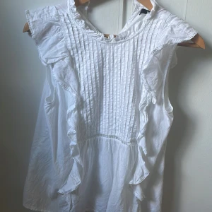 Vit blus med volang och spetsdetaljer - Supersöt vit blus från bik bok med volanger längs framsidan och fina spetsdetaljer. Blusen är ärmlös och har smala veck framtill som ger en romantisk känsla. Perfekt att styla med jeans eller kjol för en fräsch look.