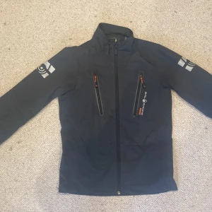 Svart vindjacka från Sail Racing - Säljer en riktigt snygg svart vindjacka från Sail Racing med snygga loggor på ärmar och rygg. Jackan har två bröstfickor med dragkedja och hög krage. Materialet är slitstarkt och lätt, perfekt för blåsiga dagar. Stilsäker design med orange detaljer på dragkedjorna.
