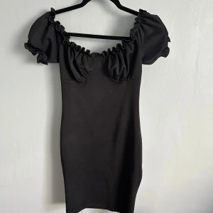 Svart kort klänning med puffärmar - Supersnygg svart kort klänning från Nelly med puffiga kortärmar och rynkade detaljer vid halsringningen. Klänningen har en figurnära passform och offshoulder-stil som ger en trendig vibe. Perfekt för dig som vill sticka ut med en enkel men cool look.