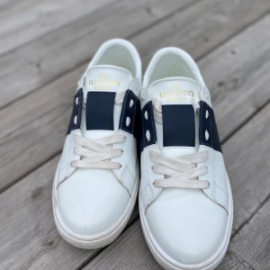Vlentino sneakers vit/blå - Snygga Valentino Garavani Open sneakers i vitt skinn med bred marinblå rem över vristen och coola nitar på hälen. Klassisk rund tå, vita snören och chunky vit sula. Perfekta för dig som gillar exklusiva och trendiga sneakers med unik design. Andvända 2 ggr, priset ej hugget i sten.