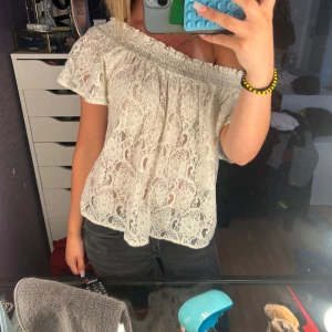 Vit spetsig offshoulder blus - Supersöt vit blus i spets med offshoulder-modell och korta ärmar. Blusen har elastisk smock upptill så den sitter snyggt både på axlarna och offshoulder. Blommigt spetsmönster och luftig passform, perfekt för varma dagar.