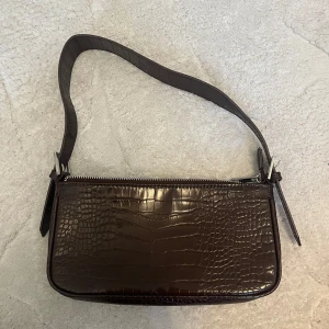 Brun axelväska från Gina Tricot - Snygg brun axelväska från Gina Tricot med croco-mönster. Har silvrig dragkedja och justerbart axelband. Använt endast 1-2 ggr