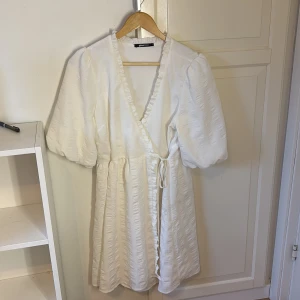 Vit omlottklänning från Gina Tricot - Superfin vit kort omlottklänning från Gina Tricot med puffiga korta ärmar och volangkant längs v-ringningen. Klänningen har knyt i midjan och lätt struktur i tyget som ger en lyxig känsla. Perfekt för sommarens alla tillfällen! Finns en liten ljusblå fläck på sidan av bysten på klänningen men inget man tänker på