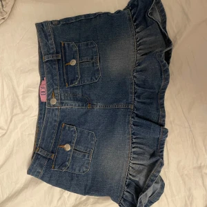 Jeanskjol med volang från DOJ - Supersöt blå jeanskjol från DOJ med volangkant nertill och två stora fickor framtill med silvriga knappar. 