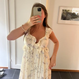 Ljusblommig långklänning med volang - Superfin långklänning i ljusbeige med blommigt mönster. Djup v-ringning med volangdetaljer, breda axelband och öppen rygg med knyt. 