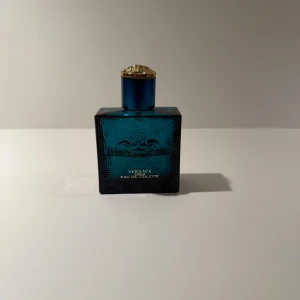 Versace Eros Eau de Toilette - Versace Eros Eau de Toilette i en stilren, fyrkantig flaska av blått glas med mönstrad yta och Medusa-huvudet i relief. Korken är blå med gulddetaljer och har Versaces ikoniska Medusa-symbol på toppen. En lyxig och modern parfymflaska som sticker ut i badrumshyllan.