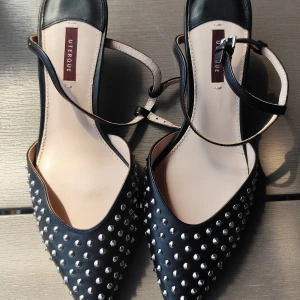 Svarta spetsiga pumps med nitar Uterque - Svarta pumps från Uterque med spetsig tå och coola silverfärgade nitar över hela skon. Skorna har en smal rem runt vristen och är tillverkade i skinn. Perfekta för dig som vill sticka ut med en edgy och trendig look.