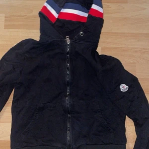 Svart hoodie från Moncler - Svart hoodiejacka från Moncler med dragkedja framtill och klassisk logga på ärmen. Huva med snygga ränder i rött, vitt och blått ger en sportig vibe. Perfekt för dig som gillar streetstyle och vill ha något bekvämt men ändå stilrent.